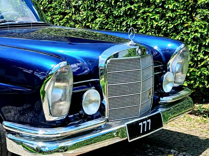 Sedã clássico azul dos anos 1960, com grade e para-choques cromados, diante de uma cerca-viva