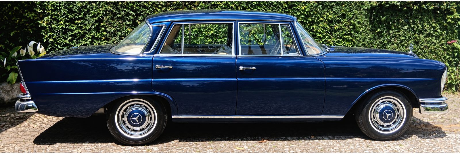 Sedã clássico azul dos anos 1960 visto de lado, com rodas de aro cromado, diante de uma cerca-viva